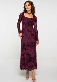 Largo vestido ajustado en terciopelo burdeos profundo con un patrón floral. Presenta largas mangas transparentes y un escote cuadrado. Acentuado con zapatos de tacón con tiras.
