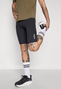 Schwarze Sportshorts mit perforierten Einsätzen, kombiniert mit weißen Knöchelsocken mit schwarzen Streifen und schwarzen Laufschuhen.