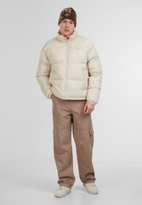 Pufu jaka off-white krāsā ar augstu apkakli un rāvējslēdzēju, apvienota ar beige krāsas tērpu biksēm un baltajām sporta apaviem. Brūns cepure ar rakstu.