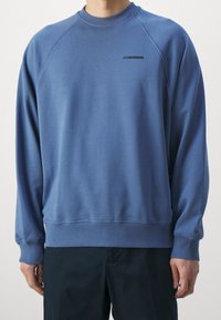 Blå crewneck-tröja med raglanärmar, gjord av mjukt tyg. Har en liten svart logga på bröstet samt ribbade muddar och fåll.