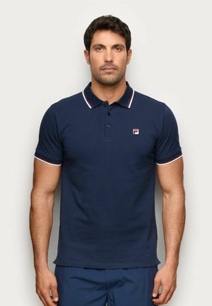 Hombre con camiseta polo azul marino de manga corta con ribete blanco y rojo en el cuello y las mangas, de pie frente a un fondo liso y claro.