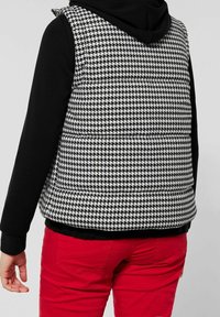 Gepolsterte Weste in einem schwarz-weißen Hahnentrittmuster, mit Kapuze und seitlichen Einsätzen aus schwarzem Stoff; getragen über roten Hosen.