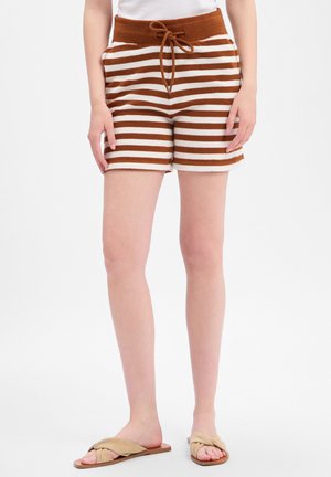 Frau trägt braune und weiße gestreifte Strickshorts mit Kordelzug und beige flache Sandalen und steht vor einem einfarbigen Hintergrund.