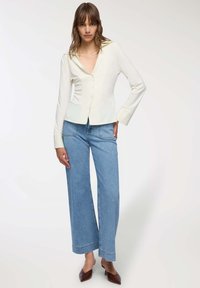 Donna che indossa una camicia bianca a maniche lunghe con bottoni, jeans larghi blu chiaro e tacchi a punta marroni mentre si trova contro uno sfondo bianco.