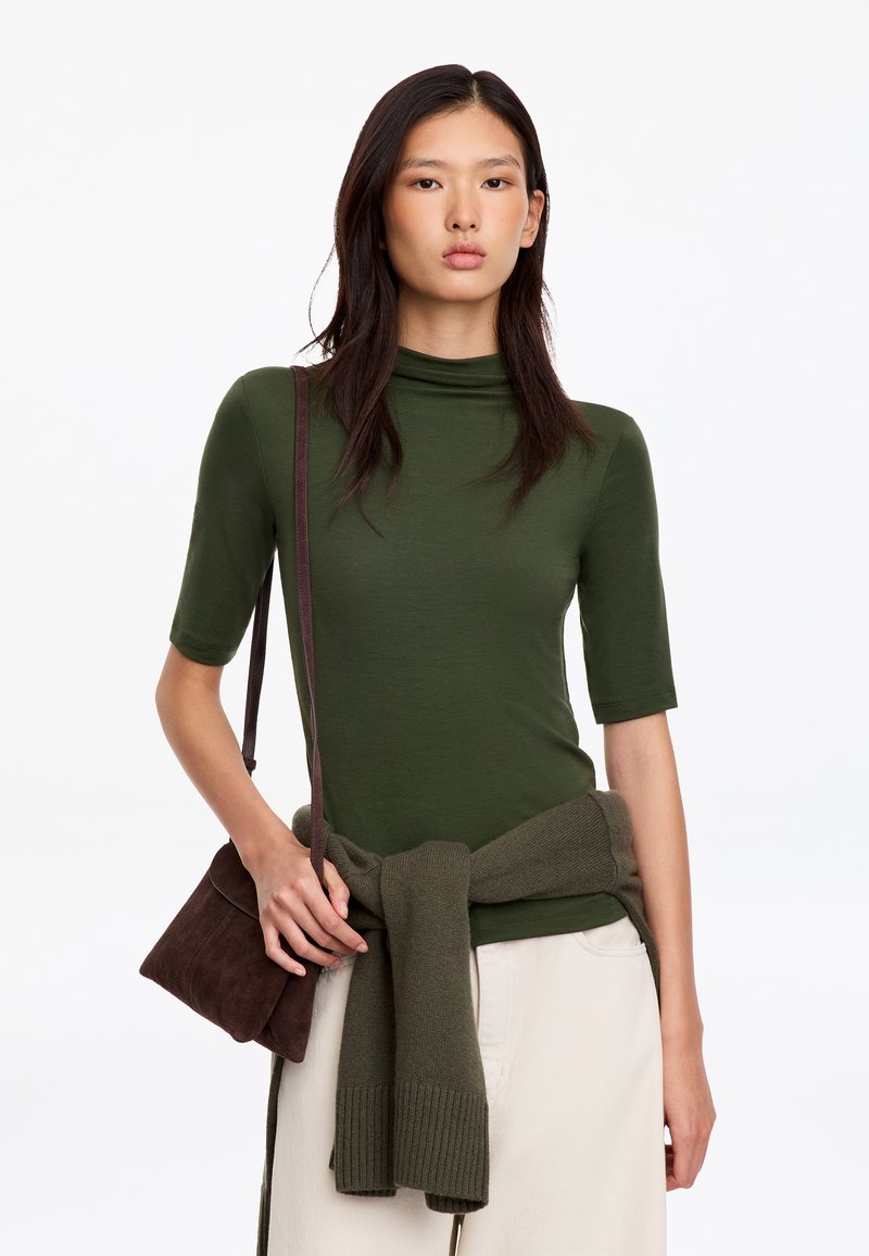 Jeune femme portant un haut vert à col montant, un pantalon crème, un pull vert noué autour de la taille et un sac à épaule marron.