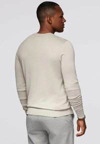 Maglione lavorato a maglia beige chiaro con colletto a coste e maniche lunghe aderenti, indossato con pantaloni grigi chiari, mostrando una texture liscia.