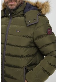 Olivengrønn polstret jakke med hette av faux pels, to-farget design, glidelås-lukking og brodert lapp på venstre erme.