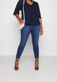 Marinebluse mit strukturierter Oberfläche, Rüschenausschnitt und Knöpfen. Kombiniert mit dunkelblauen Skinny Jeans und schwarzen Riemchenschuhen.