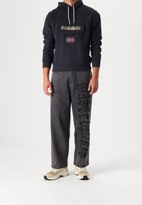 Mężczyzna w czarnej bluzie Napapijri z norweską flagą oraz szarych dżinsach z czarnym nadrukiem na prawym nogawce, w beżowych sneakersach.