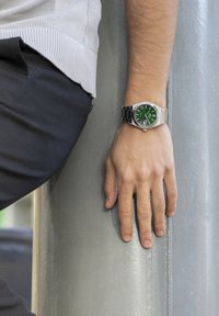 Silver wristwatch with a green dial, stainless steel bezel, and black metal band. Hand resting on a gray pole.