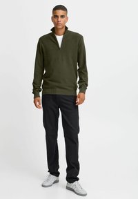 Olivgrüner Reißverschluss-Pullover mit gerippter Textur, langen Ärmeln und einem taillierten Bund. Kombiniert mit schwarzen Hosen und grauen Turnschuhen.