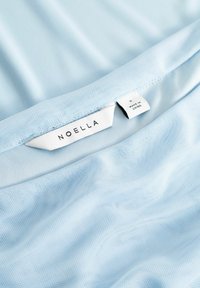 Hellblaues Mesh-Gewebe mit glatter Textur, versehen mit einem Etikett mit der Aufschrift "Noella" und einem Größenschild "S", nahe dem Saum angebracht.