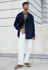 Abrigo navy de corte cruzado sobre una camisa beige, combinado con pantalones blancos y zapatos marrones tipo slip-on. El abrigo presenta grandes botones y un cuello.
