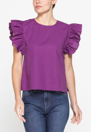 Donna con una canotta viola senza maniche con grandi dettagli arricciati sulle spalle e jeans blu, in piedi davanti a uno sfondo bianco uniforme.