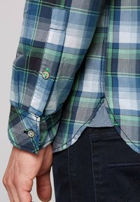 Camicia a quadri blu e verde con maniche lunghe, caratterizzata da polsini con bottoni e orlo curvo. La trama del tessuto sembra liscia e morbida.