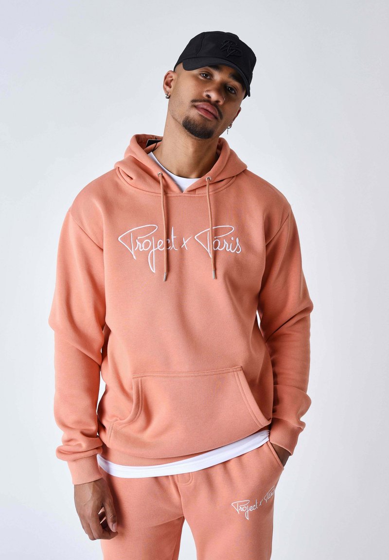 Sudadera con capucha color melocotón claro con texto de logotipo blanco, capucha ajustable con cordón, bolsillo delantero y mangas con puños; combinada con pantalones deportivos a juego.