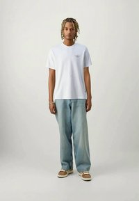 Λευκό βαμβακερό t-shirt με κοντά μανίκια και μικρή εκτυπωμένη σχέδιο, σε συνδυασμό με ανοιχτό μπλε, φαρδιά τζιν παντελόνια και καφέ αθλητικά παπούτσια.