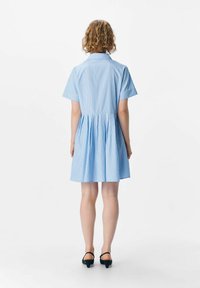 Robe rayée bleu clair avec des manches courtes, un col et un design de jupe plissée, fabriquée dans un tissu lisse. Chaussures à talons noires portées.