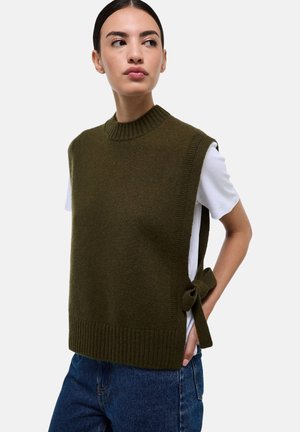 Haut en tricot sans manches de couleur vert foncé avec un col côtelé et un détail de nouage sur le côté, associé à une chemise blanche et un jean bleu.