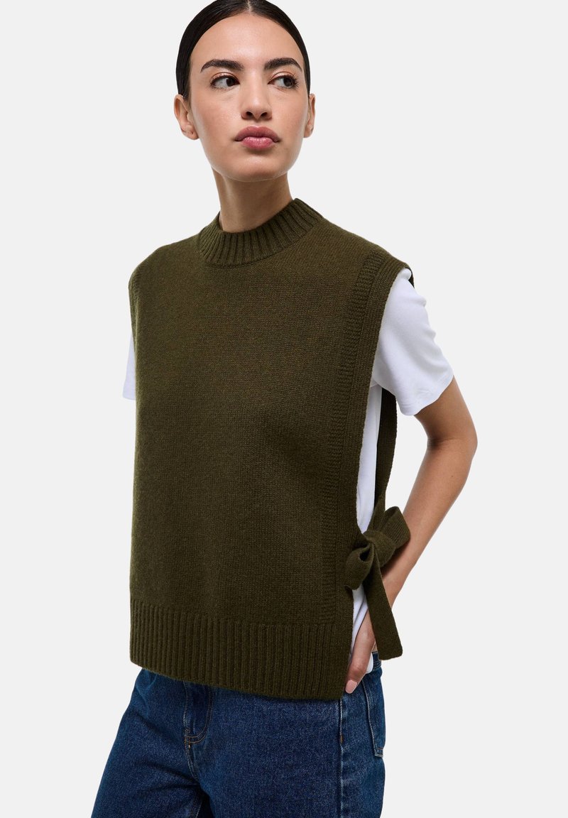 Haut en tricot sans manches de couleur vert foncé avec un col côtelé et un détail de nouage sur le côté, associé à une chemise blanche et un jean bleu.