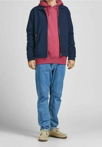 Tmavě modrá bomber bunda přes vínovou mikinu s kapucí, doplněná světle modrými džíny a béžovými botami. Má zapínání na zip a volný střih.