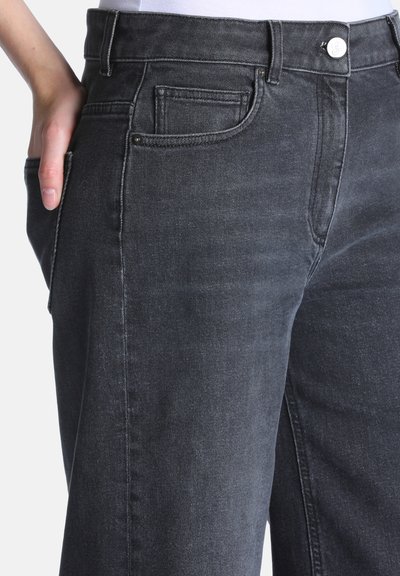Gros plan d'une personne portant un jean gris foncé, la main dans la poche arrière, sur fond blanc.