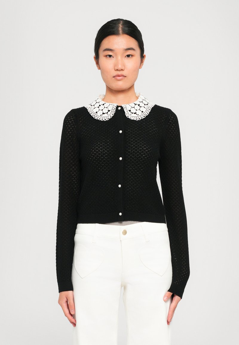 Black knitted cardigan with a white lace Peter Pan collar, featuring a button front and long sleeves, paired with white pants.