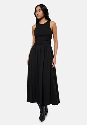Kvinde iført sort ærmeløs maxikjole med tætsiddende korset og udvidet nederdel, kombineret med sorte spidsede støvler, stående mod hvid baggrund.