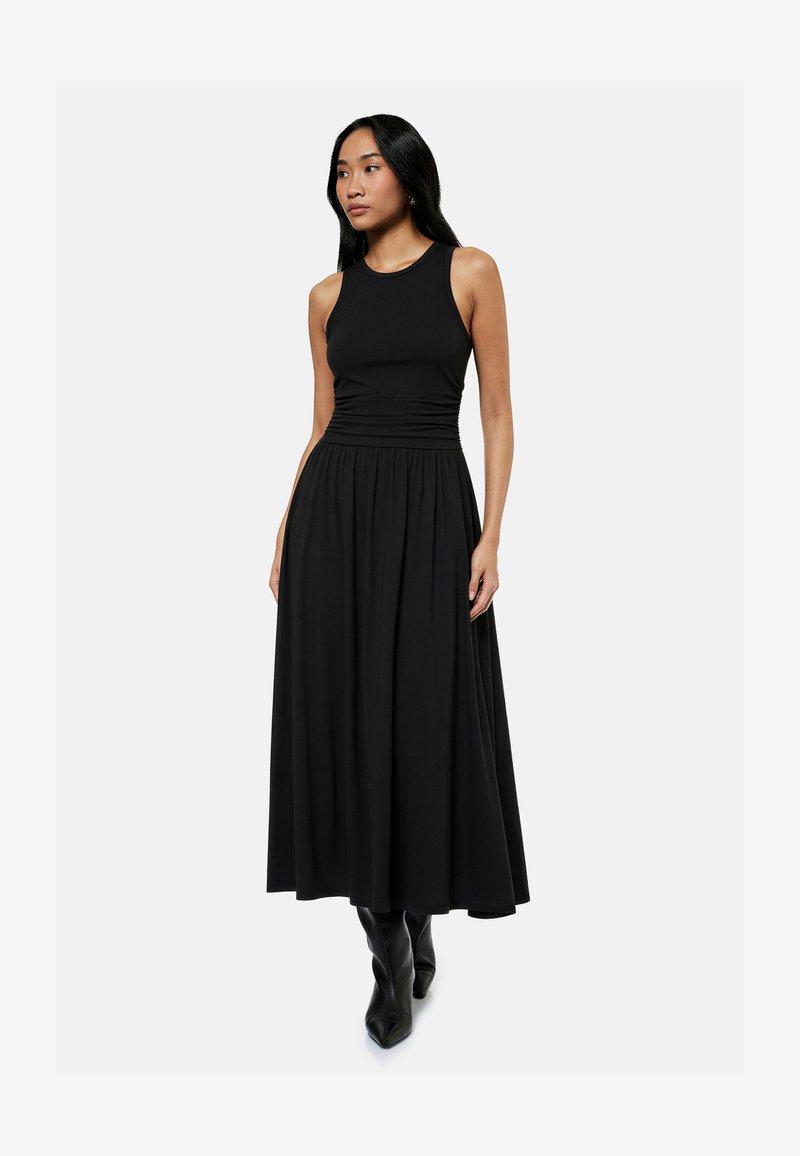 Femme portant une robe maxi noire sans manches avec un corsage ajusté et une jupe évasée, associée à des bottes noires à bout pointu, debout devant un fond blanc.