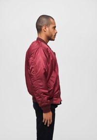 Veste bomber bourgogne avec une texture lisse, des poignets et un ourlet côtelés, des poches latérales et une fermeture éclair, présentée sur un mannequin masculin de profil.