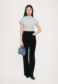 Femeie îmbrăcată cu un top scurt de culoare gri deschis, pantaloni negri cu talie înaltă, pantofi negri, ținând în mână o poșetă mică albastră, stând pe un fundal alb.