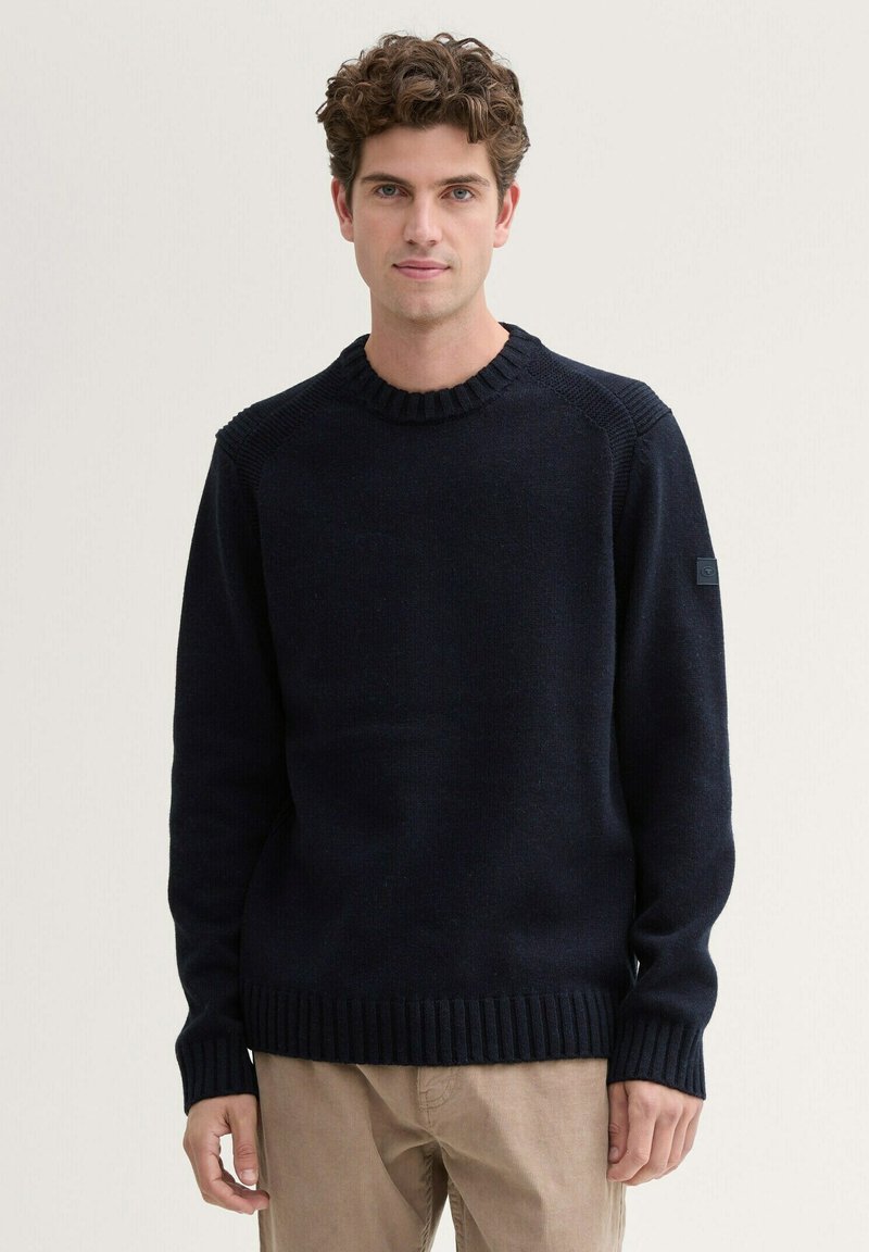 Pull en maille bleu marine avec un col et des poignets côtelés, présentant un motif texturé sur les épaules et une petite étiquette logo sur la manche.