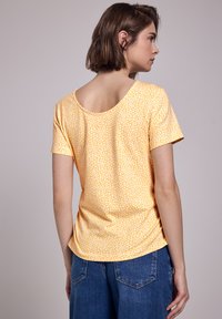 Top amarillo de manga corta con un patrón floral, escote redondo en la espalda y ajuste holgado, combinado con jeans azules.