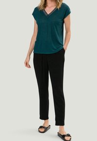 Haut en teal à manches courtes avec un col en V et des détails en dentelle, associé à un pantalon noir et des sandales à glissière noires, avec une coupe décontractée.