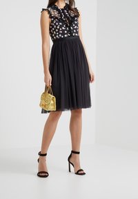 Vestido negro floral con escote rizado, superposición transparente y falda de tul. Combinado con tacones negros de tiras y un pequeño bolso dorado.