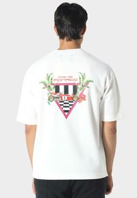Hvid oversized t-shirt med et farverigt grafisk design på ryggen, som inkluderer palmeblade, et ternet mønster og et racingmotiv.