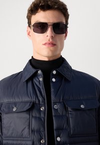 Mornarska puffer jakna z gumbi na snap in dvema sprednjima žepoma. Nosimo jo preko črnega puloverja z visokim ovratnikom, ima gladko, sijajno teksturo. Sončna očala so vključena.