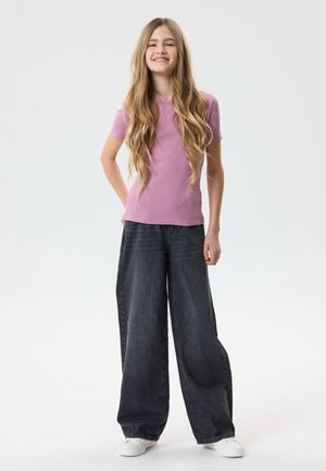 Teen girl with long blonde hair wearing a pink short-sleeve shirt, loose black jeans, and white sneakers stands smiling against a white background.