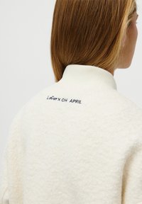 Valkoinen fleece-huppari, jossa on ribbipanta, takana navynsininen brodeeraus, jossa lukee "Lefer x OH APRIL". Pehmeä kangas.