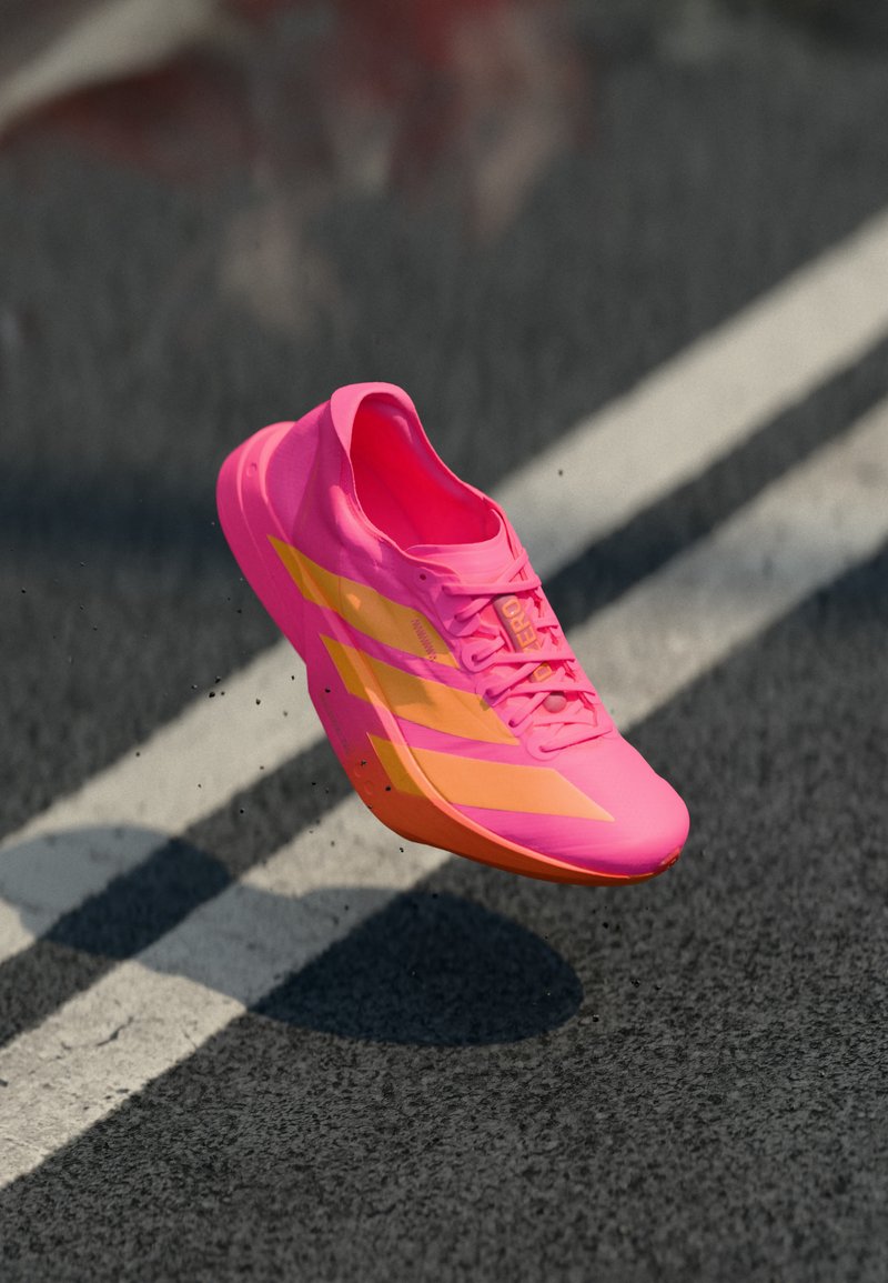 Leuchtend pinker Laufschuh mit orangefarbenen Streifen und Schnürsenkeln, schwebt über Asphalt mit weißen Fahrbahnmarkierungen.