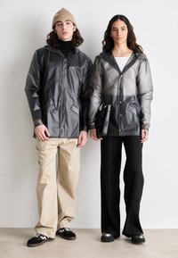 Deux personnes se tiennent côte à côte. L'une porte une veste noire brillante et imperméable avec des poches ; l'autre porte une veste transparente de couleur gris clair.
