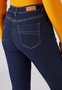 Jeans de mezclilla azul oscuro con una cintura alta, que presentan detalles cosidos, dos bolsillos traseros y una etiqueta de cuero marrón en la cinturilla.