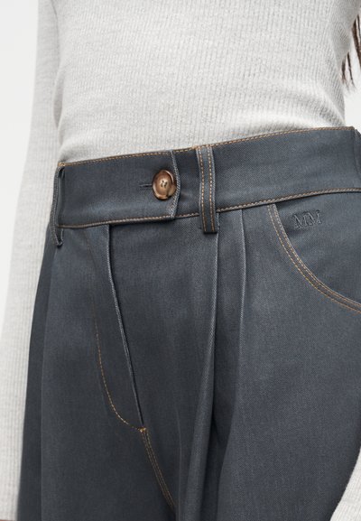 Κοντινό πλάνο σκούρων γκρι παντελονιών με ψηλή μέση, καφέ ραφές, κλείσιμο με κουμπιά και κεντημένο λογότυπο "MM", φορεμένα με ανοιχτό γκρι μπλουζάκι με ριπ υφή.