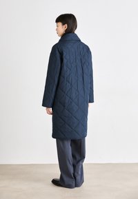 Manteau matelassé bleu marine avec col montant, manches longues et coupe droite. Présente un motif en diamant et arrive au milieu de la cuisse.