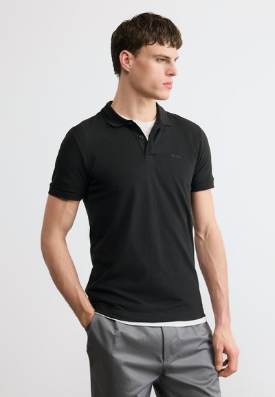 Homme portant un polo noir à manches courtes et un pantalon gris, debout avec une main dans la poche, regardant sur le côté contre un fond uni.