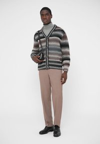 Mitmevärviline kootud kardigan hallide, mustade ja pruunide triipudega, kombineeritud beežide pükste ja mustade kingadega; käes hoiab väikest musta kotti.