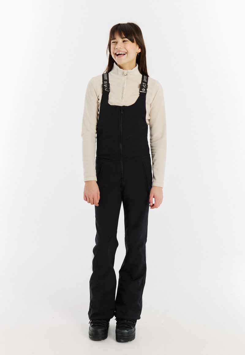 Giovane donna sorridente, che indossa una maglia a maniche lunghe beige e una salopette nera con zip e bretelle con logo, in piedi davanti a uno sfondo bianco.