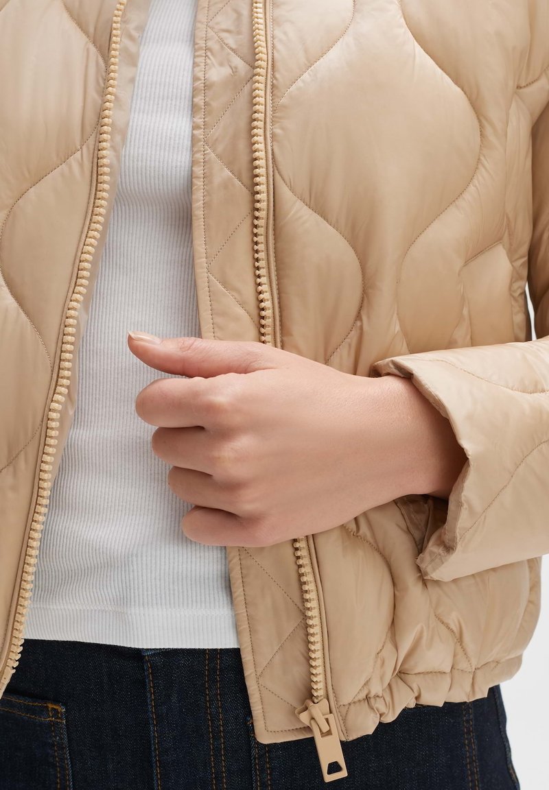 Beige gesteppte Jacke mit strukturierten Oberflächen, ausgestattet mit einem Reißverschluss und gestrickten Bündchen, getragen über einem weißen gerippten Oberteil.
