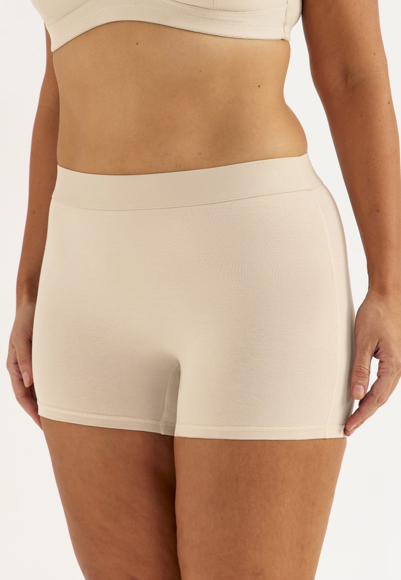 Kvinde iført beige sømløse shorts med høj talje og matchende crop top, der viser mellemgulv og øvre lår mod hvid baggrund.