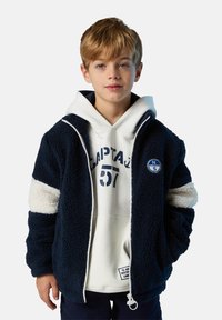 Niño rubio que lleva una chaqueta de forro polar azul marino con rayas blancas sobre un sudadera blanca con el texto "CAPTAJ 57", de pie frente a un fondo liso.